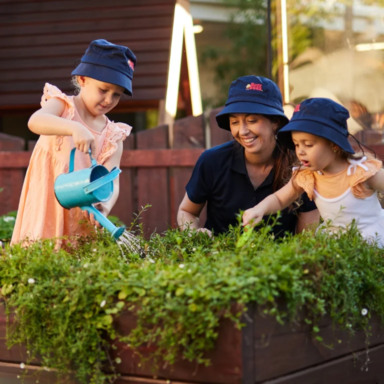 Two young girls, wearing hats and aprons, water plants in a garden bed while a woman kneels beside them, smiling and assisting. It's a delightful moment of early learning and childcare in nature's classroom.