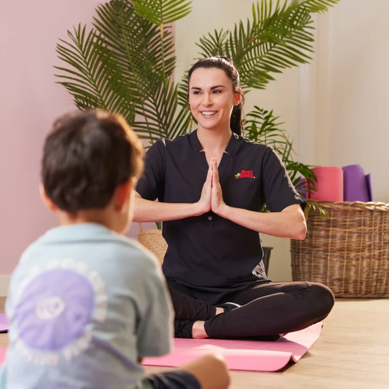 A person in a black outfit sits cross-legged on a pink mat with hands in prayer position, smiling at a child seated across from them. The cheerful setting resembles a preschool, with indoor plants and colorful yoga mats in the background.