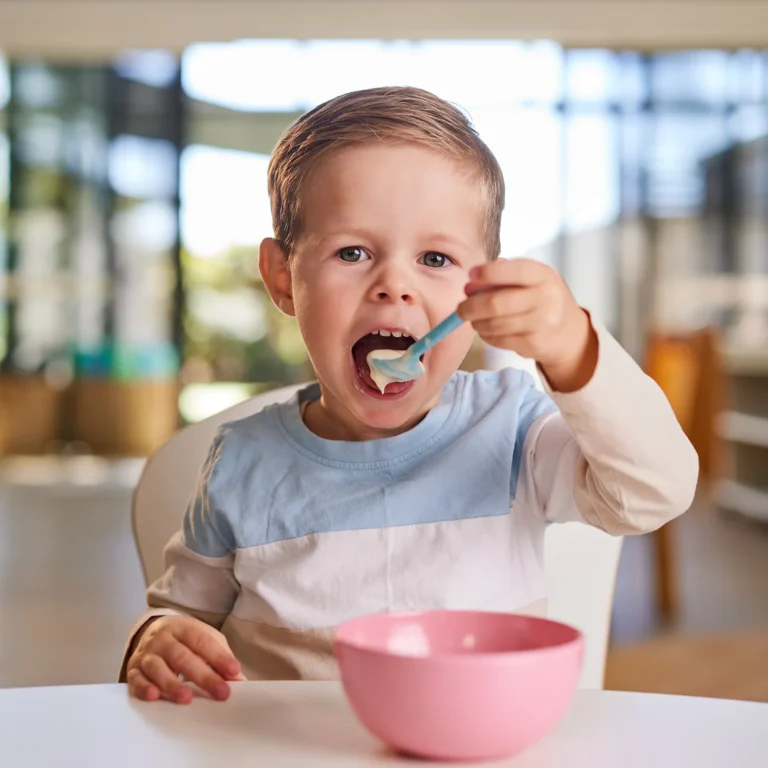 A young child sits at a table in a bright room, enjoying a meal from a pink bowl with a blue spoon. The scene exudes the warmth and care of quality childcare, highlighting the essence of early learning.