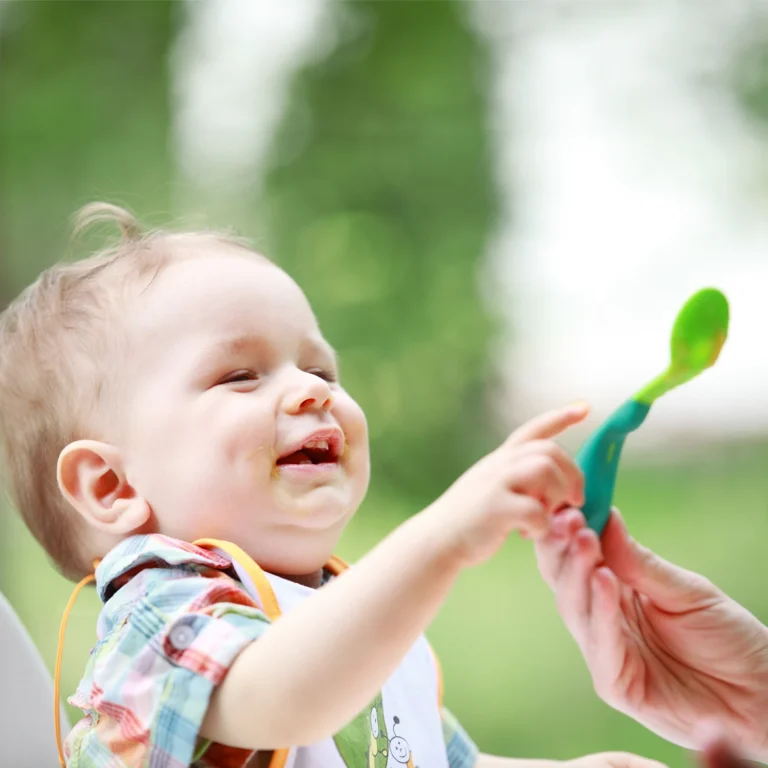 A baby wearing a bib reaches for a spoon while sitting outdoors at day care. The baby, with food smeared on its mouth, is being fed by an adult.