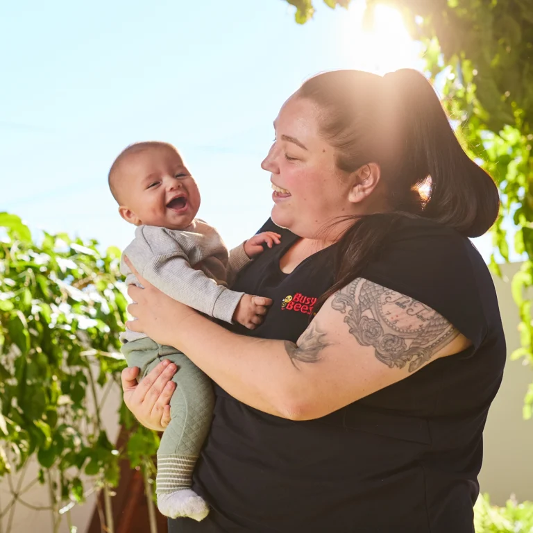 A woman holds a smiling baby outdoors at a preschool. Sunlight filters through the trees around them. The woman has a tattoo on her arm and is wearing a black shirt with a red logo.