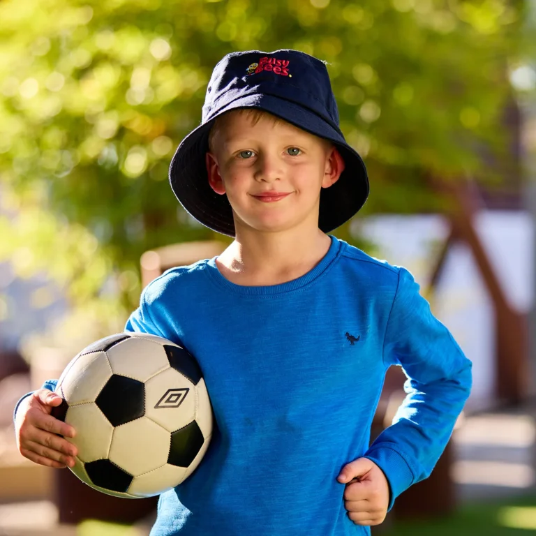 A young boy wearing a blue shirt and bucket hat, likely from preschool, holds a soccer ball under his arm while standing outdoors.