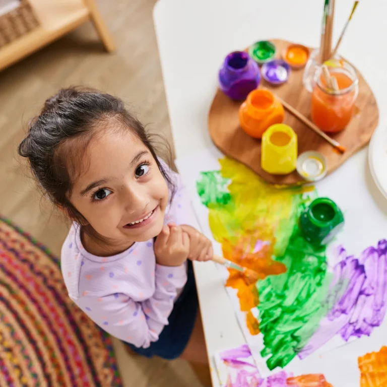 A young child with a top-knot hairstyle is smiling while painting with various colors on a white paper at a preschool table. Paint jars and brushes are visible nearby.