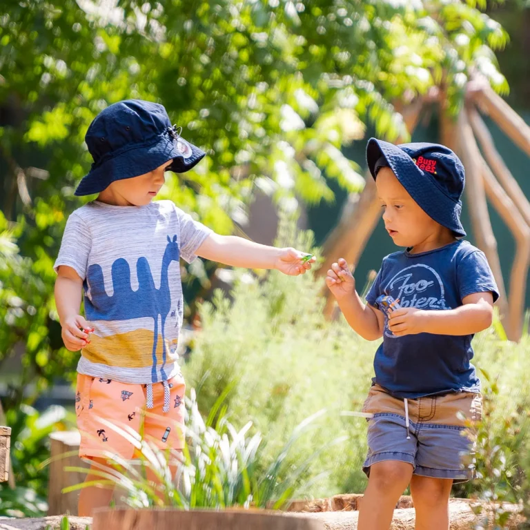 Two young children wearing sun hats are outdoors in a garden at their daycare. One child is handing a small green item to the other, fostering early learning through play. Both are casually dressed and surrounded by greenery.