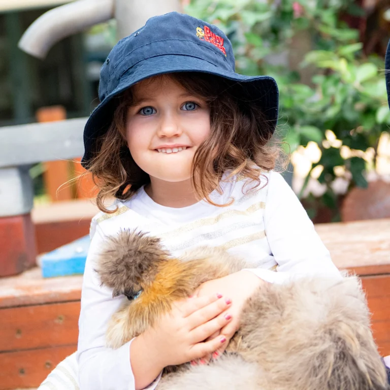 A young girl from kindergarten wearing a navy bucket hat, holding a fluffy chicken, and smiling with her mouth slightly open.