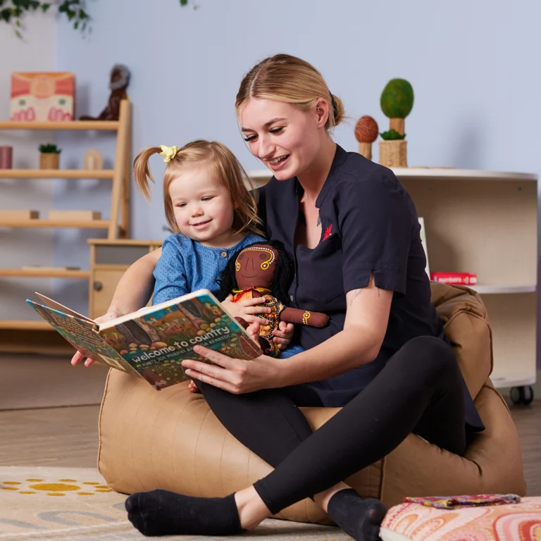 A woman and a young girl are sitting on a pouf, reading a colorful book together in a cozy room with shelves of toys, perfect for early learning.