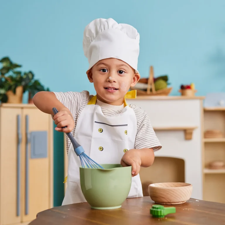 A young child, part of a vibrant preschool program, wears a chef's hat and apron as they mix ingredients with a whisk in a green bowl within a lively kitchen setting.