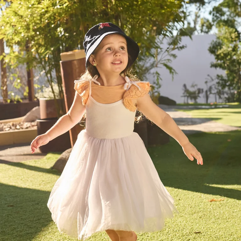 A young girl in a white dress and black hat walks outside on a sunny day, surrounded by greenery, as if she's on an adventure from her preschool.