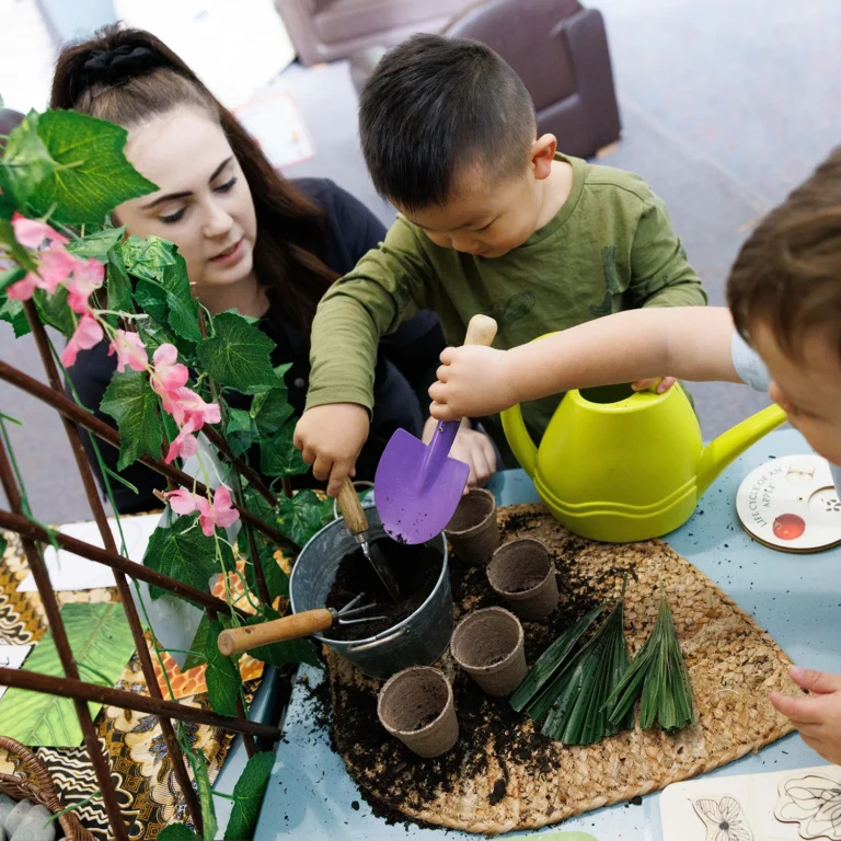 Three children are joyfully engaged in a planting activity, using gardening tools and small pots, fostering early learning. An adult from the day care is attentively assisting them.