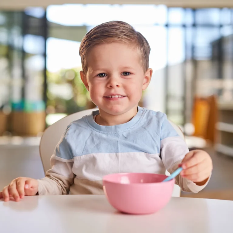 A young child with short hair is sitting at a table holding a spoon and smiling, with a pink bowl in front of him. The background shows a brightly lit room with windows, creating an inviting atmosphere perfect for early learning in kindergarten.