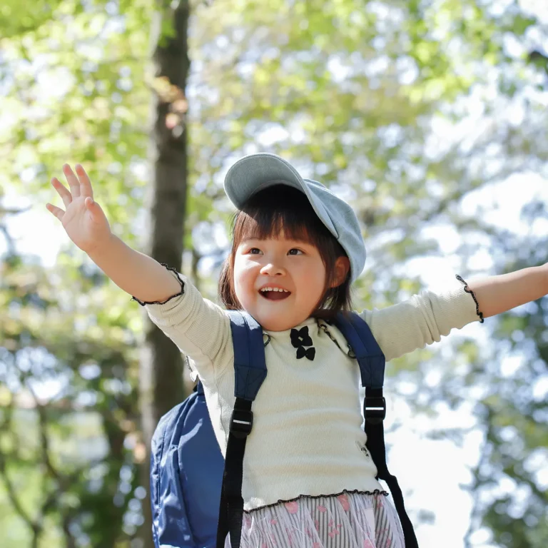 A young child wearing a cap and backpack, fresh from preschool, raises their arms enthusiastically while standing outdoors surrounded by trees.