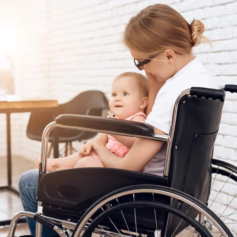 A woman in a wheelchair holds a baby on her lap. They are both smiling and sitting in a sunlit room with white brick walls, reminiscent of a cozy preschool setting.