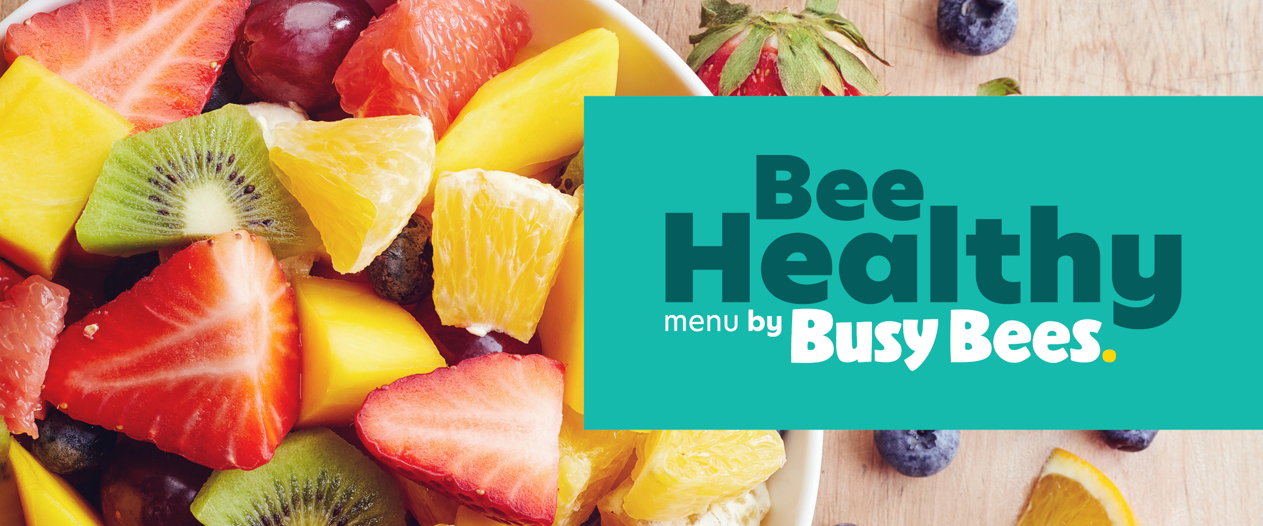 A bowl of mixed fruit, including strawberries, kiwi, mango, and oranges, beside a teal sign reading "Bee Healthy menu by Busy Bees".